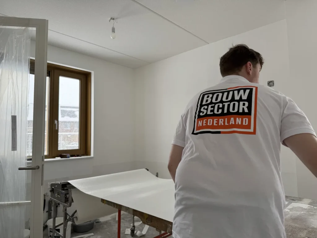 Behanger van Bouwsector Nederland rolt een baan renovlies uit over de plaktafel naast een raam met houten kozijn